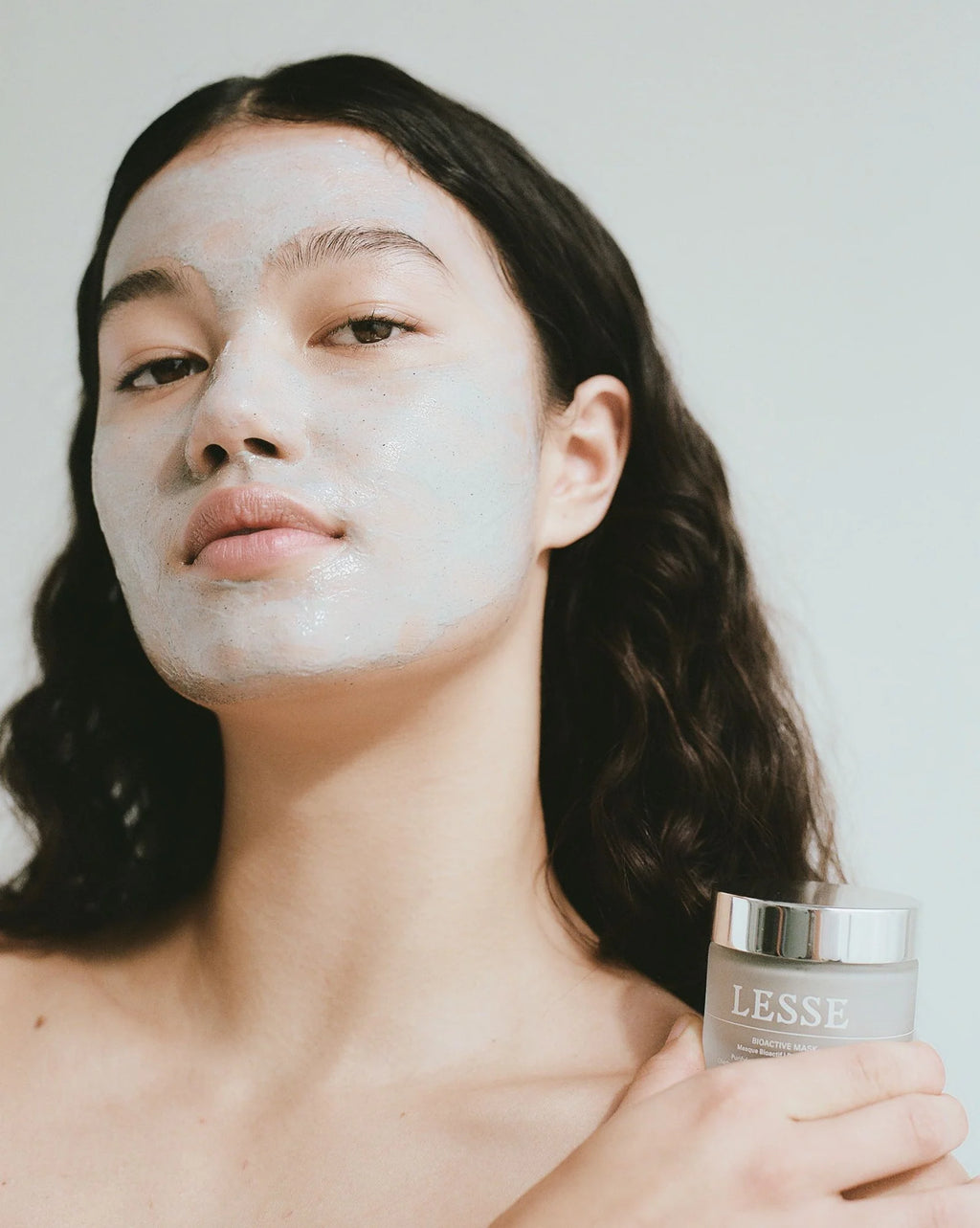 Person with a face mask holding a jar of 'LESSE' skincare product against a neutral background