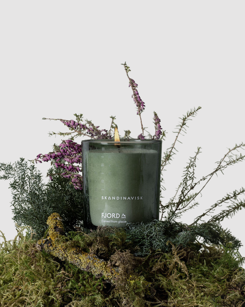 Green candle in a glass container with Brandinavik branding on a bed of moss and plants.